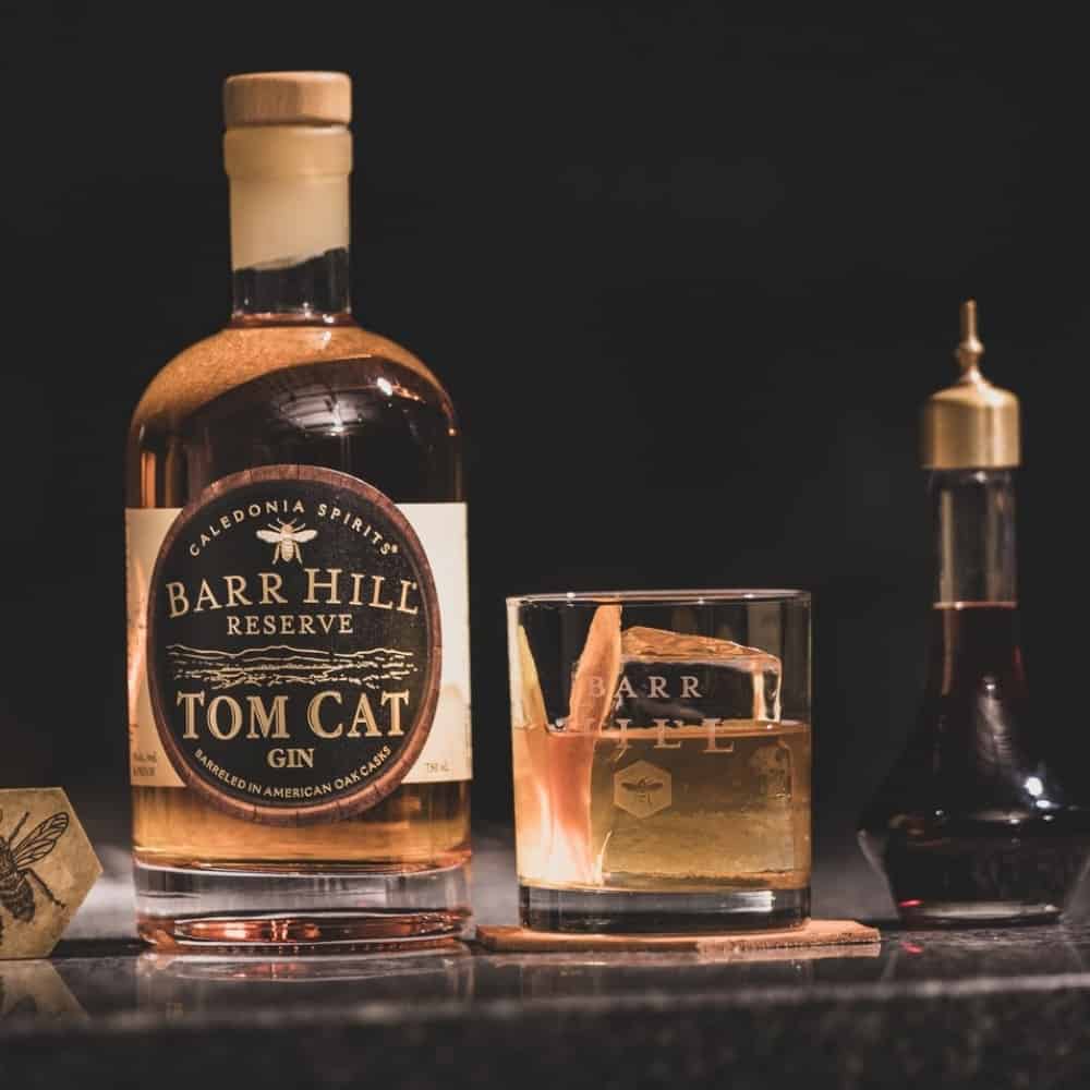 A Gin Old Fashioned in a cocktail glass next to a bottle of Barr Hill Tom Cat Gin