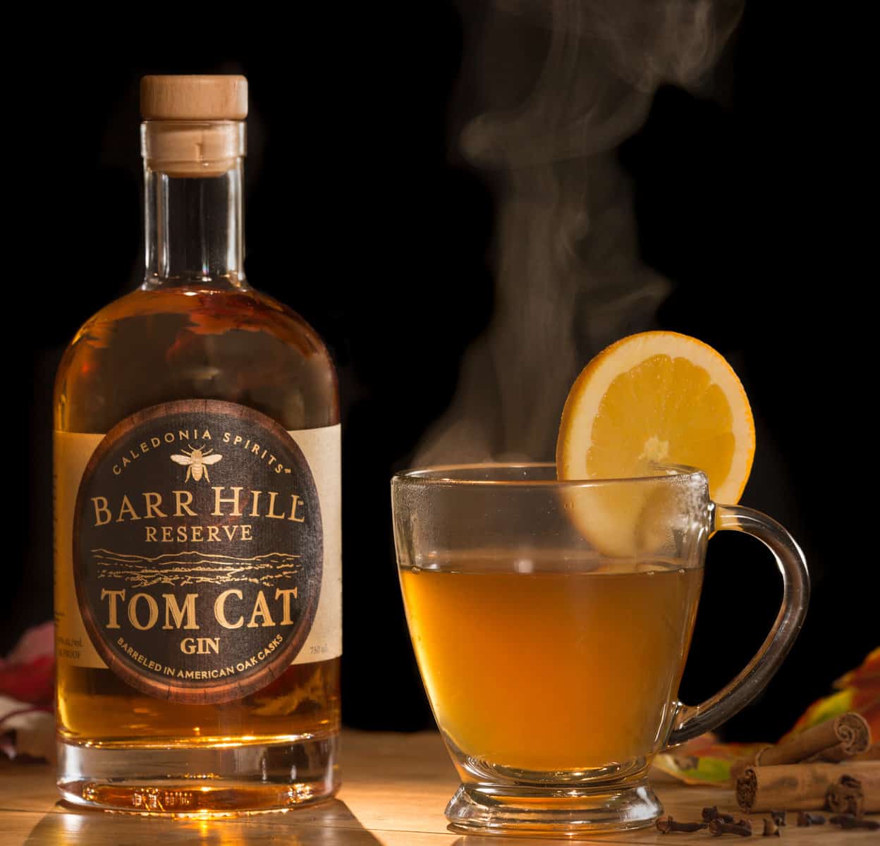 A Gin Hot Toddy with a slice of orange on the glass next to a bottle of Barr Hill Tom Cat Gin