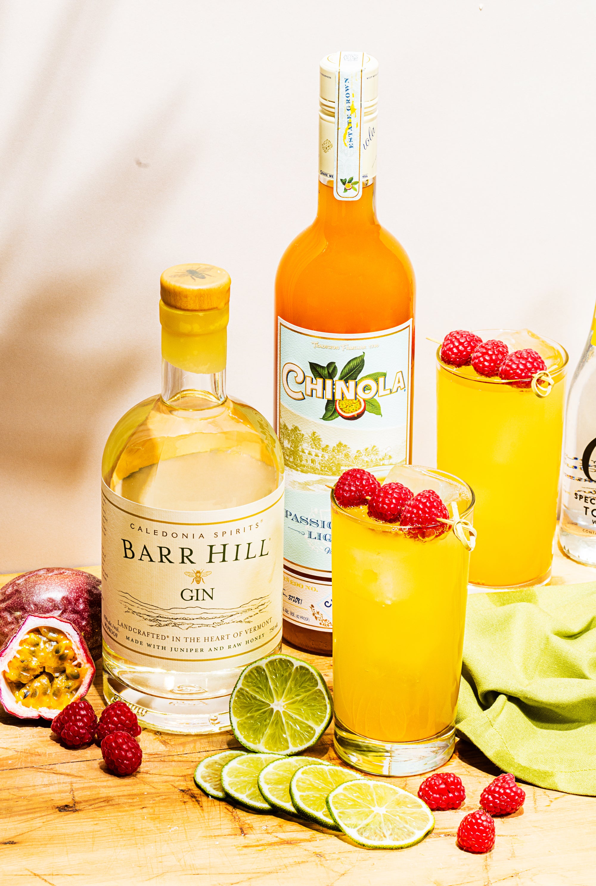 Tropic and Tonic Cocktails with bottles of Barr Hill Gin and Chinola, surrounded by slices of lime, raspberries, and passionfruit.