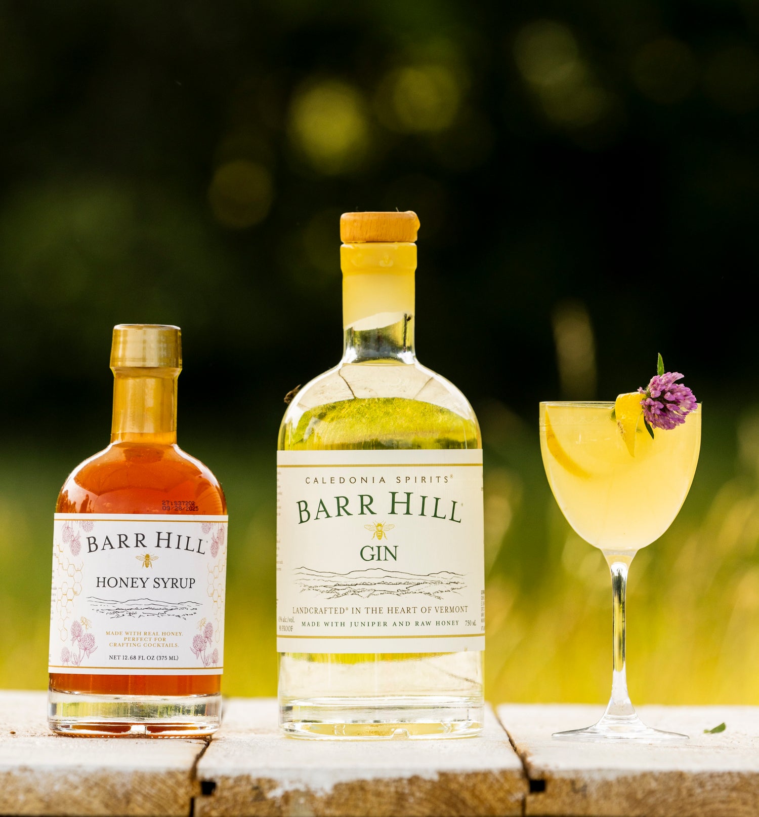 Bottles of Barr Hill Honey Syrup and Gin with a cocktail on a wooden surface outdoors.