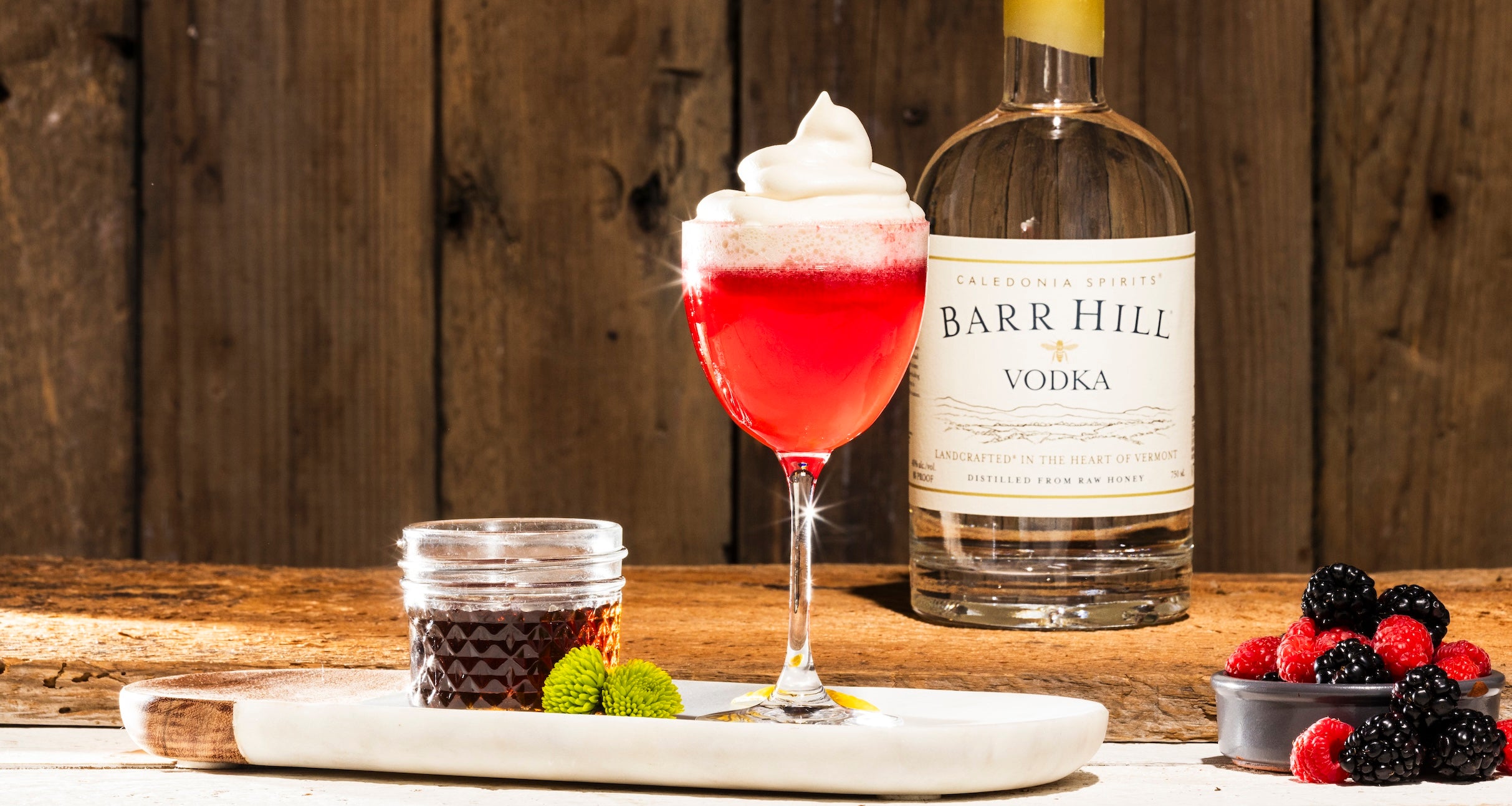 Bright red maple and berry cocktail with foam on top next to a bottle of Barr Hill Vodka, a jar of maple syrup and some blackberries and raspberries.