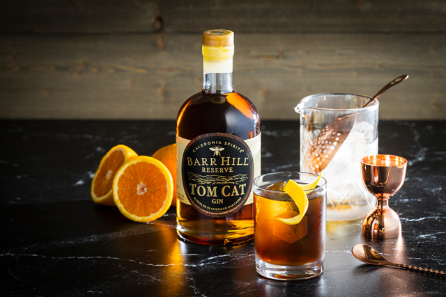 Bottle of Barr Hill Reserve Tom Cat Gin with a negroni cocktail and garnishes on a dark surface.