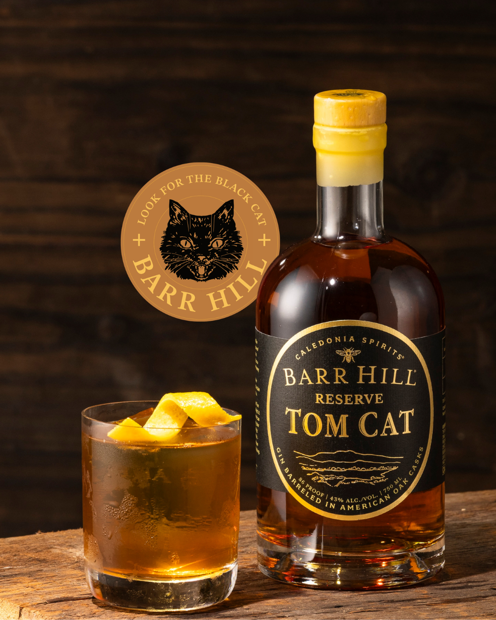 A bottle of Barr Hill Reserve Tom Cat Gin alongside a glass of amber colored Negroni cocktail with orange peel and ice, set on a rustic wooden table with a dark wood background.