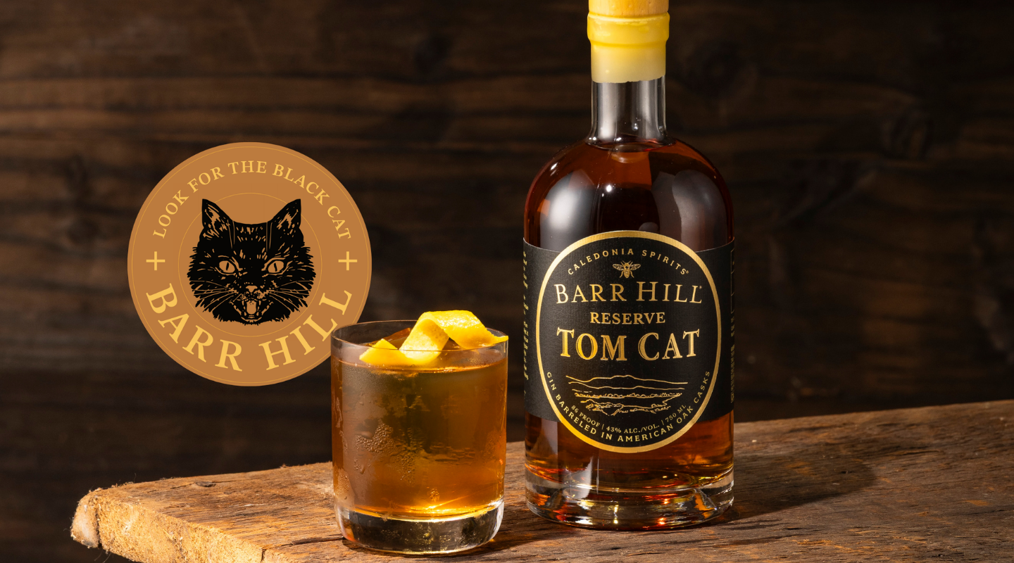 A bottle of Barr Hill Reserve Tom Cat Gin stands on a rustic wooden surface beside a rocks glass filled with an amber Negroni cocktail and orange peel garnish. A circular Barr Hill logo with a black cat illustration appears to the left against a dark wood background.