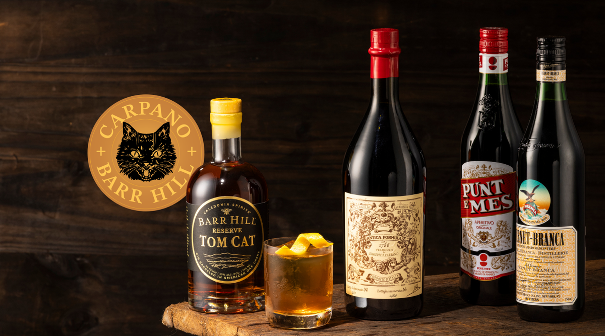 A collection of bar and cocktail ingredients arranged on a rustic wooden surface with several liquor bottles next to an amber Negroni cocktail. The lineup includes a bottle of Barr Hill Reserve Tom Cat Gin, Carpano Antica Sweet Vermouth, Punt e Mes Vermouth, and Fernet Branca.