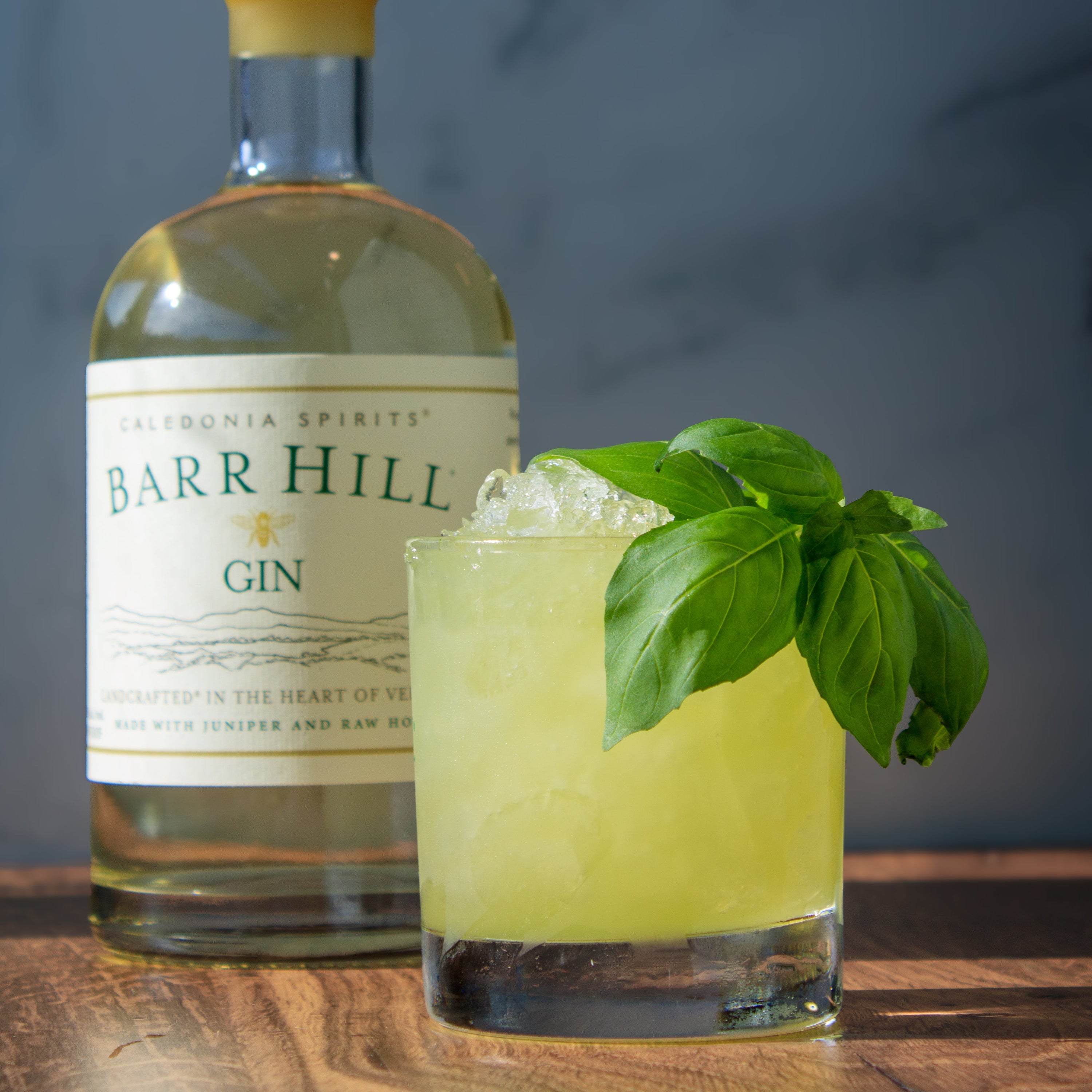 Light green cocktail in a glass with crushed ice and garnish with basil sitting on a wooden table next to a bottle of Barr Hill Gin.