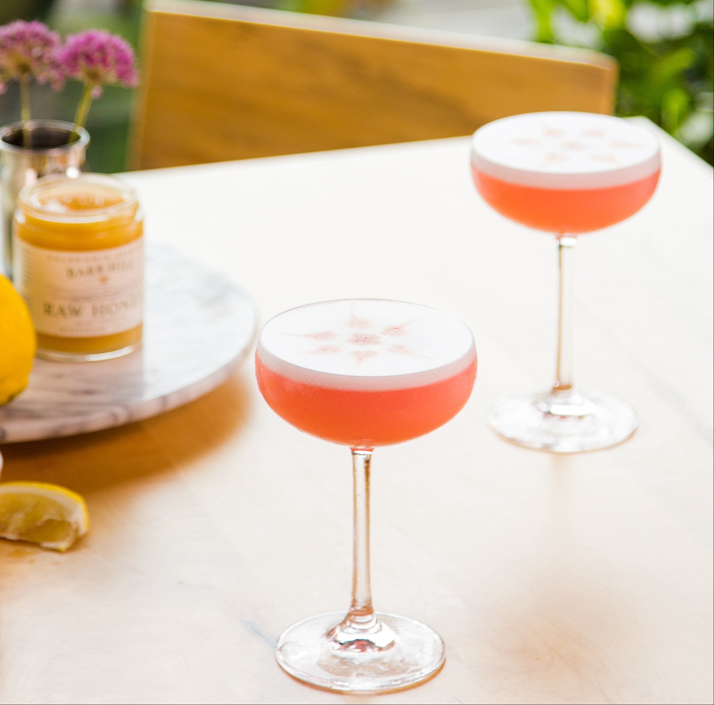 Two pink cocktails with foam on a table with lemons and plants in the background