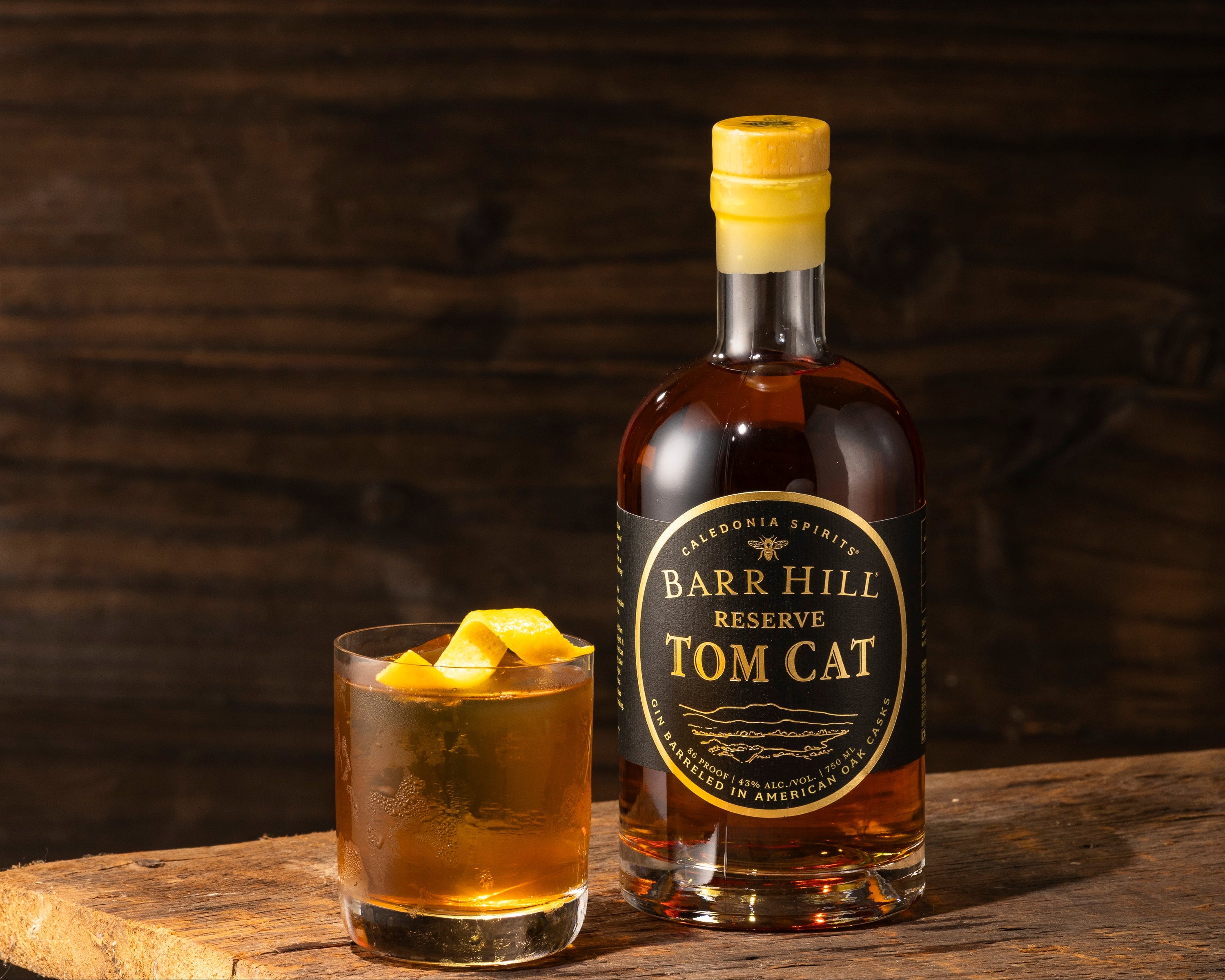 A bottle of Barr Hill Reserve Tom Cat Gin stands on a rustic wooden surface next to a rocks glass filled with a cocktail garnished with an orange peel, set against a dark wood background.