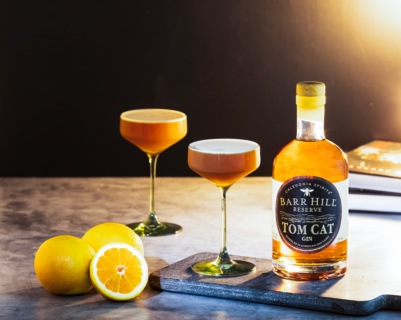 Bottle of Barr Hill Reserve Tom Cat Gin with two hive noir negroni cocktails and lemons on a wooden surface.